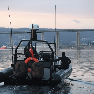 A person in a wetsuit stands at the stern of a rigid inflatable boat (RIB) equipped with navigation equipment, antennas, and safety gear, on a calm waterway at dusk with a large bridge and distant shoreline visible in the background.