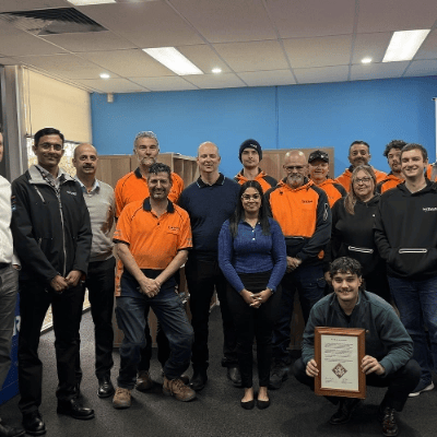 A group of approximately fifteen people, some wearing orange high-visibility workwear and others in dark uniforms or casual business attire, are gathered indoors in front of a blue wall, with one person in the front row crouching and holding a framed certificate or award.
