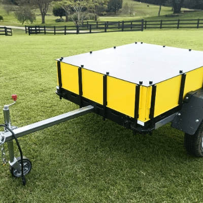 A small utility trailer with yellow side panels and a white top cover is parked on a grass lawn, with a wooden fence and trees visible in the background.