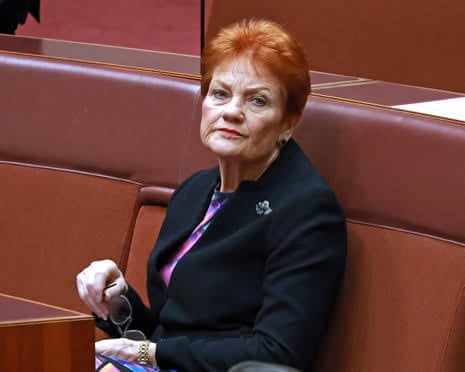 A person with short red hair, wearing a black blazer with a brooch and a pink top, is seated in a red-upholstered chamber seat, holding a pair of glasses.