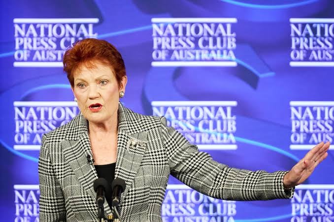 A woman with short red hair, wearing a houndstooth blazer, speaks at a podium with microphones in front of a repeated "National Press Club of Australia" branded backdrop.