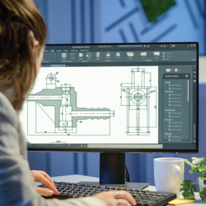 A person is seated at a desk working on a keyboard while viewing detailed CAD engineering drawings displayed on a desktop monitor in an indoor office setting.