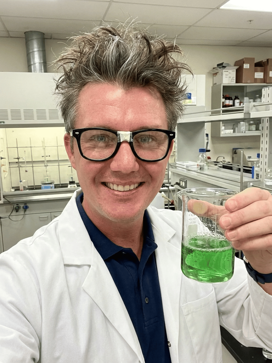 A man in a white lab coat and black-rimmed glasses with disheveled hair holds up a beaker filled with bright green liquid inside a laboratory.