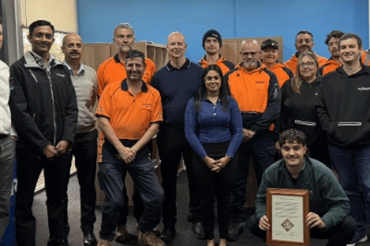 A group of approximately fifteen people, some wearing orange high-visibility workwear and others in dark uniforms or casual business attire, are gathered indoors in front of a blue wall, with one person in the front row crouching and holding a framed certificate or award.