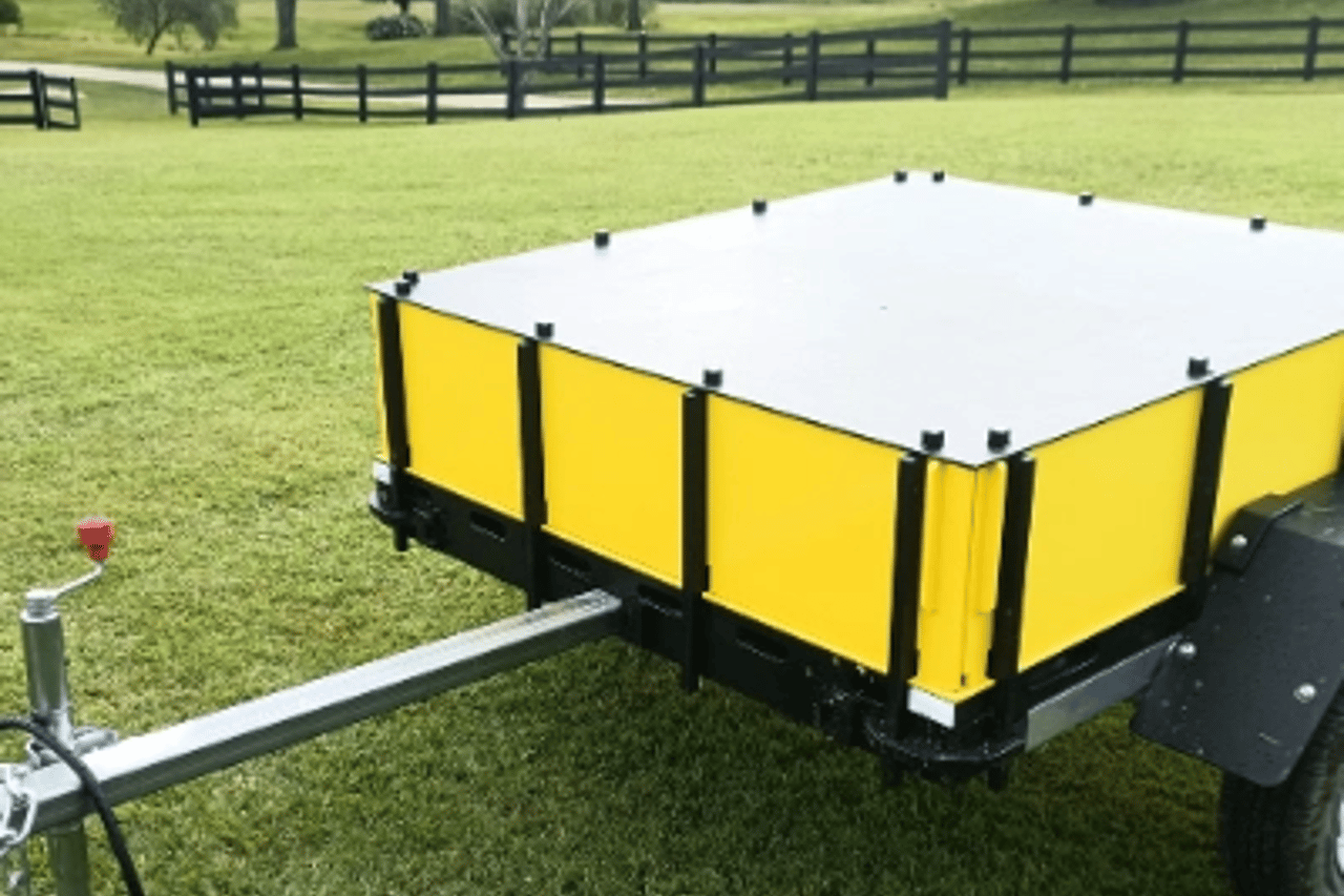 A small utility trailer with yellow side panels and a white top cover is parked on a grass lawn, with a wooden fence and trees visible in the background.